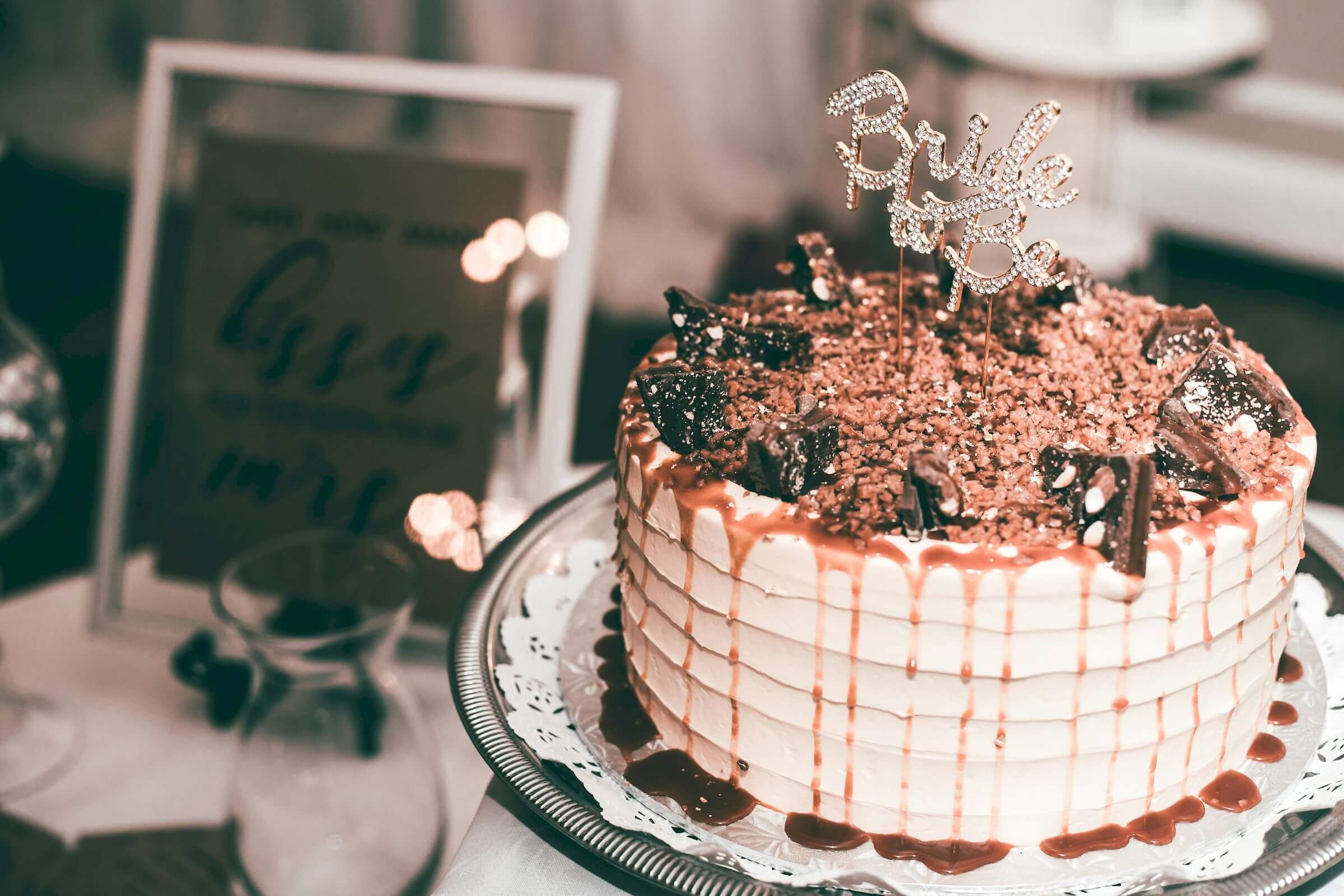Layered cake with caramel drizzle and chocolate decorations on a white lace doily, topped with a "Bride" sign. Nearby, a framed sign is slightly blurred in the background.
