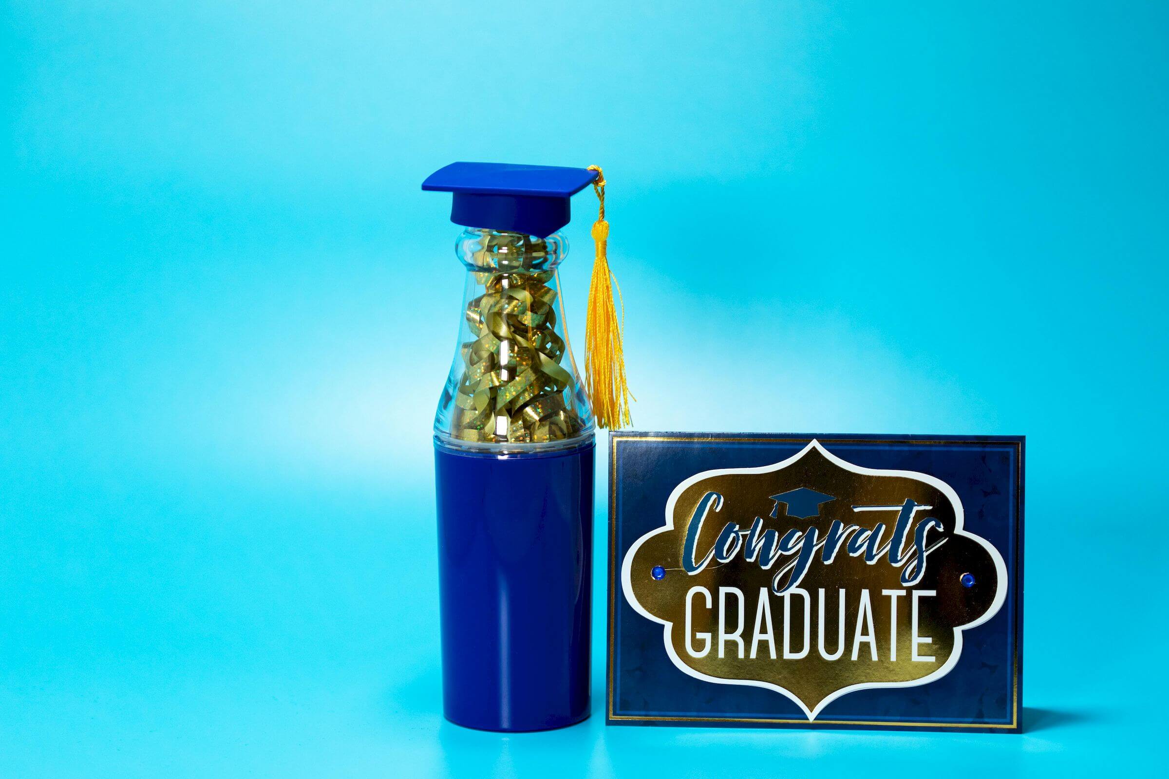 Graduation-themed bottle with a mortarboard cap beside a card reading "Congrats Graduate" on a blue background.