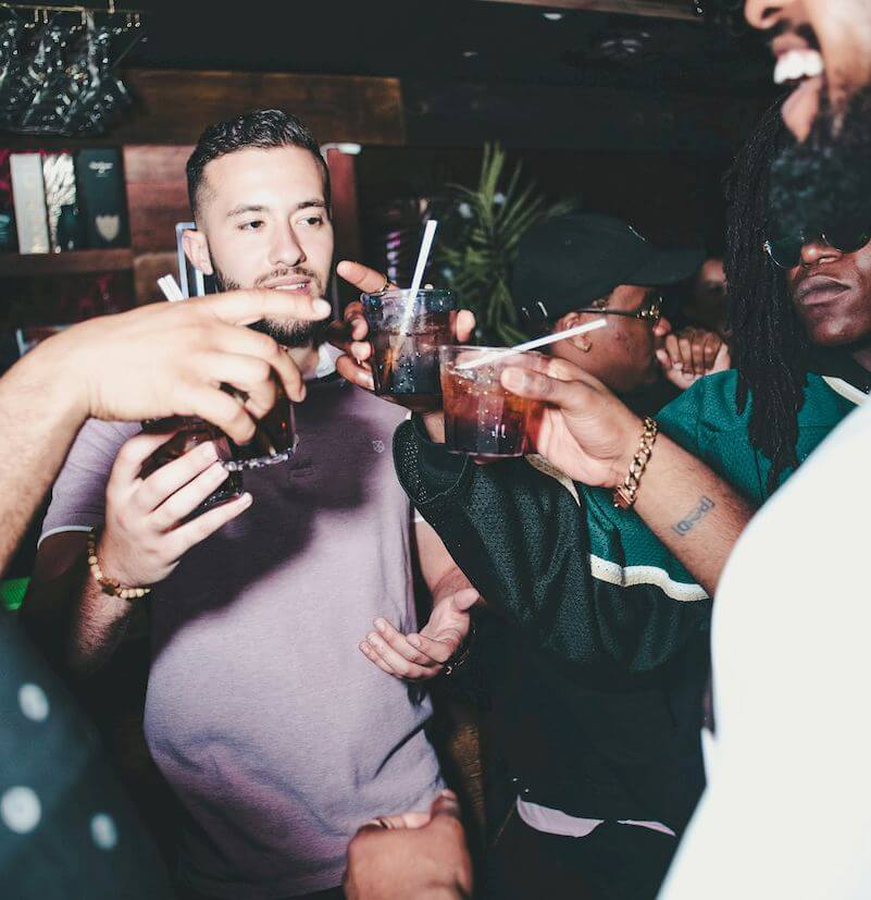 A group of people clinking glasses in a dimly lit setting, with one person in a purple shirt holding a drink.